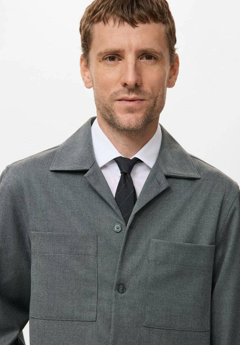 Grijze textuurjacket met een kraag, voorzien van twee voorzakken en een knoopsluiting, gedragen over een wit overhemd en een zwarte strik.