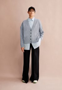 Grauer Strickcardigan mit schwarzen Knöpfen, über einem hellblauen Hemd getragen. Kombiniert mit schwarzen, weit geschnittenen Hosen und weißen Sneakers mit grünen Akzenten.