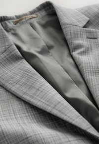 Blazer de cuadros grises con tejido texturizado, que cuenta con un forro interior suave. Los detalles notables incluyen solapas afiladas y un bolsillo en el pecho.