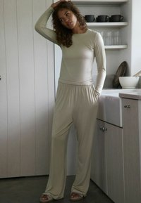 Kvinde i beige langærmet top og vide bukser står i køkkenet, den ene hånd i lommen, den anden hånd rører hendes krøllede hår.