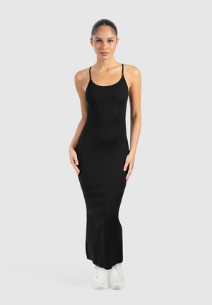 Schwarzes, ärmelloses, geripptes Maxikleid mit figurbetontem Schnitt und dünnen Trägern. Kombiniert mit weißen Sneakers, präsentiert vor einem schlichten Hintergrund.