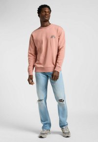 Różowy sweter z grafiką słońca, luźny krój. Dopasowany do jasnoniebieskich zniszczonych dżinsów i szarych sneakersów. Delikatne ściągacze przy mankietach i kołnierzu.