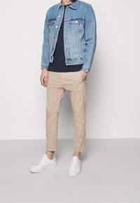 Lichtblauwe spijkerjas, marineblauw T-shirt, beige chinos en witte sneakers. De jas heeft twee borstzakken en een licht rafelige zoom.