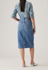 Kvinna som bär en jeansjacka och en högmidjad, knälång jeanskjol, står med ena handen på höft, vita klackskor.