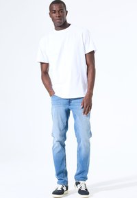 Hombre de pie con las manos en los bolsillos, vistiendo una camiseta blanca lisa, jeans de mezclilla azul claro y zapatillas en blanco y negro, sobre un fondo blanco.