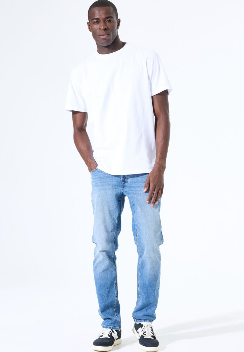 Hombre de pie con las manos en los bolsillos, vistiendo una camiseta blanca lisa, jeans de mezclilla azul claro y zapatillas en blanco y negro, sobre un fondo blanco.