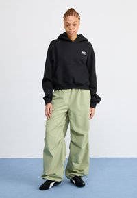 Svart hettegenser med frontlomme, parret med løse, lysegrønne bukser. Modellen har på seg svarte joggesko; minimal merkevare på hettegenseren.