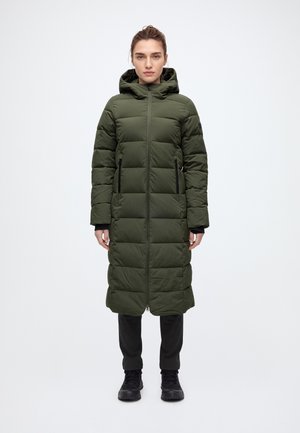 Une femme porte un long manteau matelassé à capuche vert olive, doté d’une fermeture éclair sur le devant et de poches, associé à un pantalon noir et à des bottes.