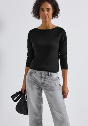 Haut noir à manches longues avec un col large, associé à un jean large gris clair. La personne tient un petit sac noir. Design simple, textures minimales.