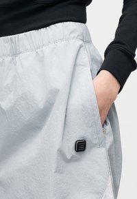 Άτομο που φοράει ανοιχτό γκρι παντελόνι με ελαστική μέση, χέρι στην πλαϊνή τσέπη και μαύρο τετράγωνο λογότυπο κοντά στη ραφή της τσέπης.