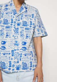 T-shirt med kort ärm gjord av lättviktigt tyg, med ett blått och vitt tryckt mönster som har trädgårdsillustrationer och text. Knappstängning framtill.