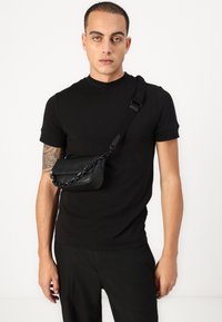 Černá crossbody taška vyrobená z kůže s zakřiveným tvarem, řetízkem na rameno a magnetickým klopovým uzavřením. Nese ji osoba v černém tričku.