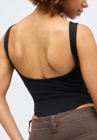 Νεαρό άτομο με σγουρά μαλλιά, που φοράει μαύρο αμάνικο τοπ και καφέ παντελόνι, απεικονίζεται από πίσω και ελαφρώς πλάγια.