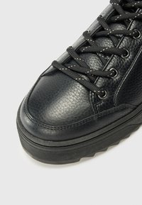 Gros plan sur une sneaker en cuir noir montrant une tige texturée, un bout rond et des lacets plats noirs passés à travers des œillets en métal sur un fond blanc.