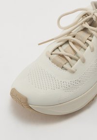 Lys beige sportsko med en teksturert overdel, snøring og en polstret såle. Har pustende mesh-deler og gummidetaljer.