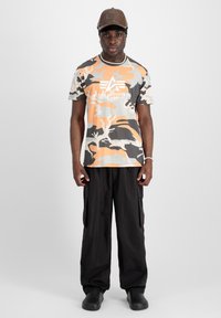 Kamouflage t-shirt i orange, grå och vit med logotyp; svarta cargobyxor; svarta slip-on skor; avslappnad keps. Tydliga färger och mönster synliga.