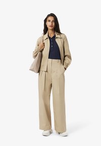 Beige jakke med elastiske manchetter over en marineblå polo t-shirt, parret med højhalset beige vidde bukser og hvide sneakers, mens der holdes en lys taske.
