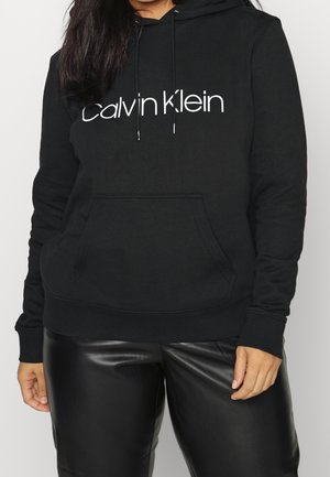 Femme portant un sweat à capuche noir Calvin Klein avec poche avant et un pantalon en cuir noir, debout devant un fond clair uni.