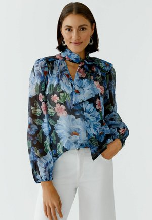 Vrouw draagt een doorschijnende blouse met grote blauwe en roze bloemen, een strikceintuur en witte broek tegen een effen achtergrond.
