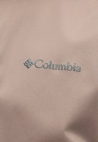 Όνομα και λογότυπο της μάρκας Columbia κεντημένα σε γκρι χρώμα πάνω σε φόντο από ανοιχτό καφέ ύφασμα.