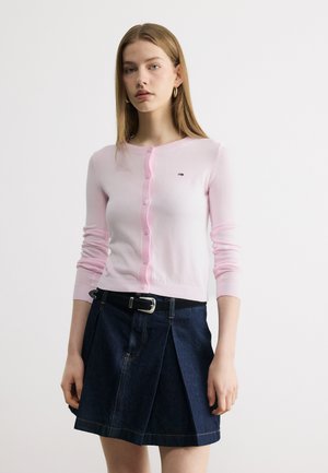 Fiatal nő világos rózsaszín gombos kardigánban és sötétkék pliszírozott farmer szoknyában fekete övvel, egy sima háttér előtt állva.