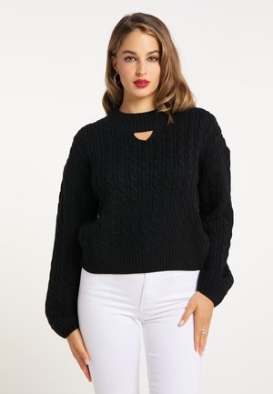 Kvinde med lyst brunt hår iført en sort kabelstrikket sweater med nøglehul-detalje ved halsen og hvide bukser, stående mod en ensfarvet baggrund.