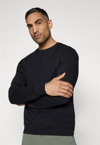 Μαύρο φούτερ από μαλακό ύφασμα, με στρογγυλή λαιμόκοψη και μακριά μανίκια, φορεμένο με τα χέρια σταυρωμένα σε ουδέτερη φόντο.