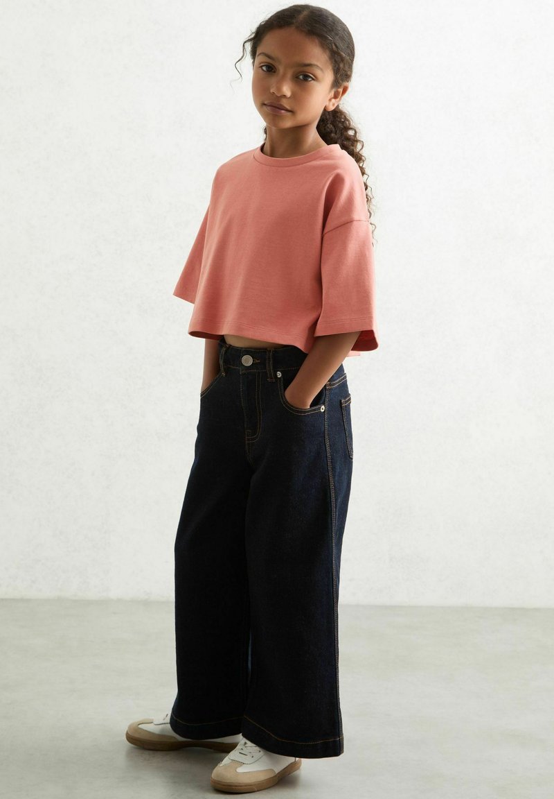 Lyserød cropped t-shirt med korte ærmer, parret med brede, mørke denim jeans og hvide sneakers, som har synlig syning og lommer.