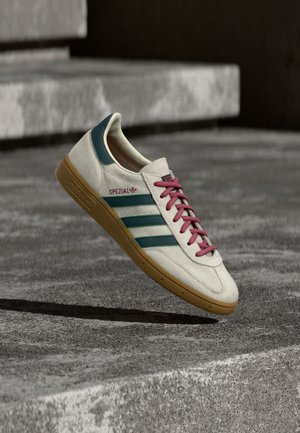 Λευκό αθλητικό παπούτσι Adidas Spezial με πράσινες ρίγες και ροζ κορδόνια που αιωρείται πάνω από μια επιφάνεια από σκυρόδεμα με σκαλοπάτια στο φόντο.