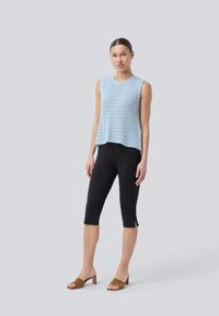 Lys blå heklede topp med bølget kant, parret med svarte capri leggings og brune åpne hæler, satt mot en nøytral bakgrunn.
