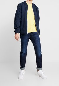 Chaqueta de bombardero azul marino, camiseta amarilla, jeans de mezclilla oscuros y zapatillas blancas. La chaqueta tiene cierre frontal y puños acanalados. Los jeans están arremangados en los tobillos.