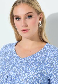 Blauw-wit luipaardprint top met een V-hals en geplooide details bij de borst. Grijze achtergrond en zilveren oorbellen maken de look compleet.