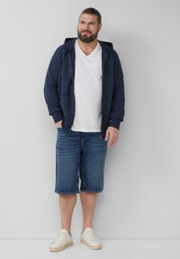 Granatowy hoodie z kieszeniami z przodu, biały t-shirt z dekoltem w serek, niebieskie szorty dżinsowe i białe sneakersy z teksturowanymi podeszwami. Prosty, casualowy strój.