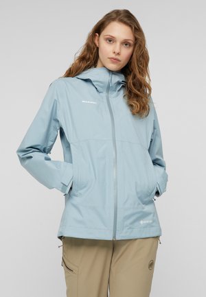 Frau trägt eine hellblaue Mammut Gore-Tex Jacke mit Händen in den Taschen und beigen Outdoor-Hosen vor einem grauen Hintergrund.
