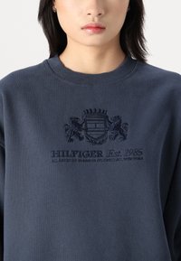Sweatshirt bleu marine en coton, avec un col rond côtelé, un logo brodé avec des lions et du texte, et une texture douce.