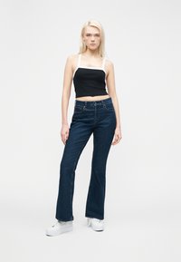 Νεαρή γυναίκα με μαύρο και λευκό κοντό τοπ χωρίς μανίκια, σκούρο μπλε ψηλόμεσα τζιν και λευκά παπούτσια πλατφόρμα, στέκεται μπροστά σε απλό φόντο.