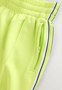 Gros plan sur un pantalon décontracté jaune fluo avec taille élastique, poche latérale zippée et finitions blanches, noires et en mesh le long de la couture latérale.
