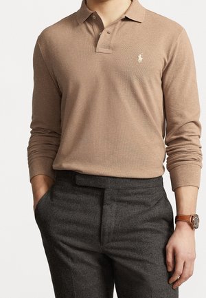 Mężczyzna w beżowej, długorękawowej koszulce polo z logo, szarych spodniach i brązowym skórzanym zegarku z srebrną tarczą, ręce w kieszeniach, na białym tle.