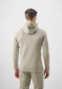 Felpa con zip beige, texture morbida, design aderente, dotata di cappuccio e tasche laterali, con accenti grigio chiaro a contrasto lungo le cuciture.