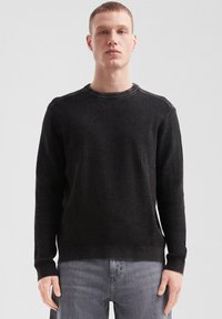 Pull en maille noire texturée avec un col rond et des manches longues, présentant des poignets et un ourlet côtelés. Porté avec un jean gris clair.