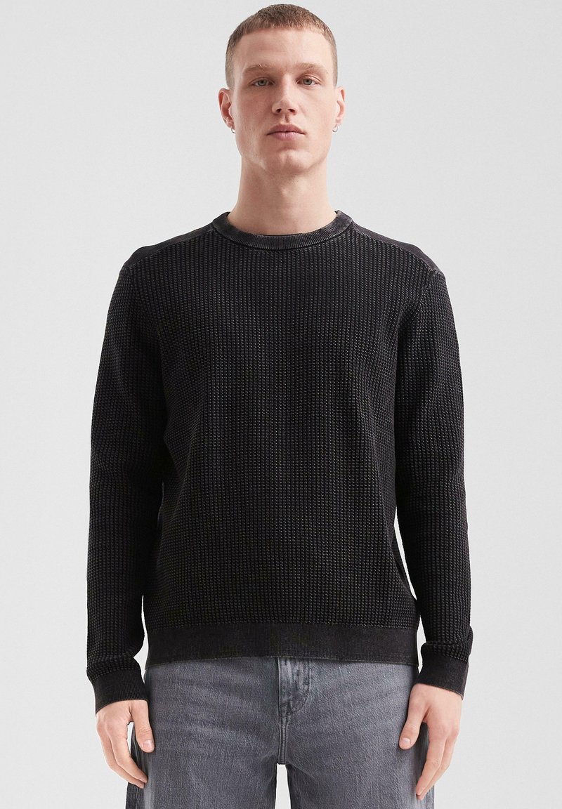 Pull en maille noire texturée avec un col rond et des manches longues, présentant des poignets et un ourlet côtelés. Porté avec un jean gris clair.