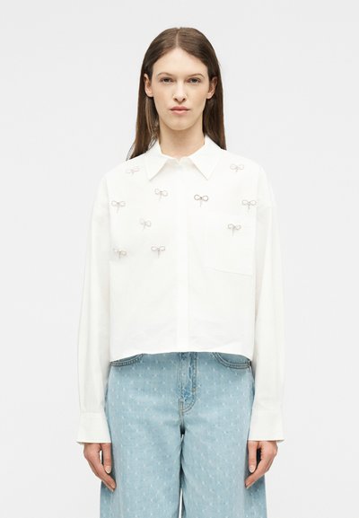 Γυναίκα που φοράει λευκό πουκάμισο με κουμπιά και μικρά κεντημένα φιόγκια, καθώς και ανοιχτό μπλε ψηλόμεσο τζιν με διακριτικά μοτίβα αστεριών.