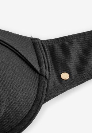 Közeli kép fekete, texturált szövet melltartó pántjáról, amelyen egy kis, kerek aranygomb található, rajta az "ALBERT TUNER" felirattal, fehér háttéren.