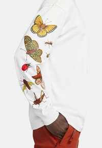 Vit långärmad skjorta med färgglada insektsillustrationer, inklusive fjärilar, nyckelpigor och gräshoppor, på vänster ärm.