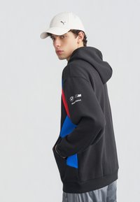 Jeune homme portant une casquette blanche Puma et un sweat à capuche BMW Motorsport noir avec des accents rouges et bleus, les mains dans la poche frontale, regardant sur le côté.