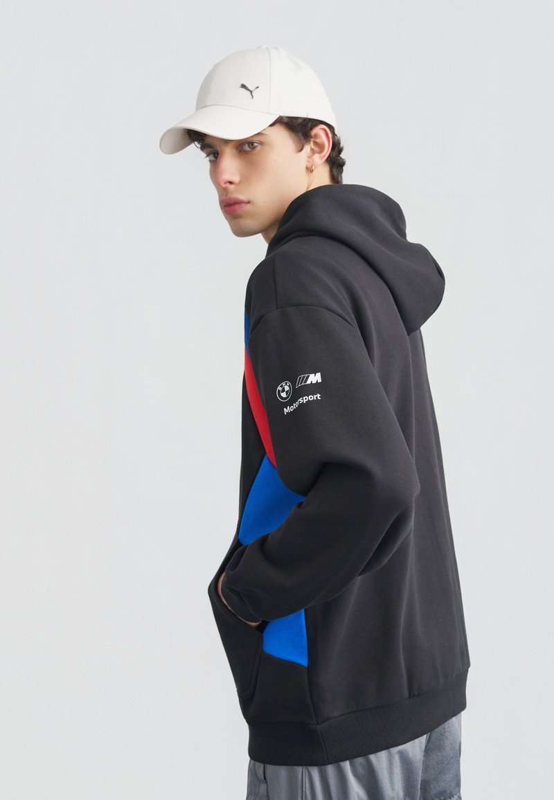 Jeune homme portant une casquette blanche Puma et un sweat à capuche BMW Motorsport noir avec des accents rouges et bleus, les mains dans la poche frontale, regardant sur le côté.