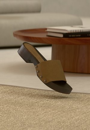 Braune Ledersandale mit niedrigem Blockabsatz, leicht über einem beigen gewebten Teppich, im Hintergrund runder Holztisch und Bücher.