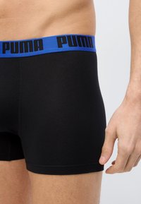 Άνδρας που φοράει μαύρα ανδρικά εσώρουχα boxer με μπλε ζώνη μέσης με το όνομα της μάρκας "Puma" σε μαύρα γράμματα, όρθιος με το χέρι σε χαλαρή θέση.