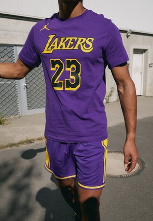 Person iført en lilla og gul Lakers basketballtrøje med nummer 23, stående udendørs nær en bygning med en metal dør.