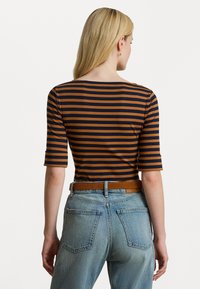 Ženska z dolgimi blond lasmi, oblečena v rjavo in črno črtasto majico z rokavi do komolca, modre kavbojke in rjav usnjen pas, obrnjena stran od kamere.